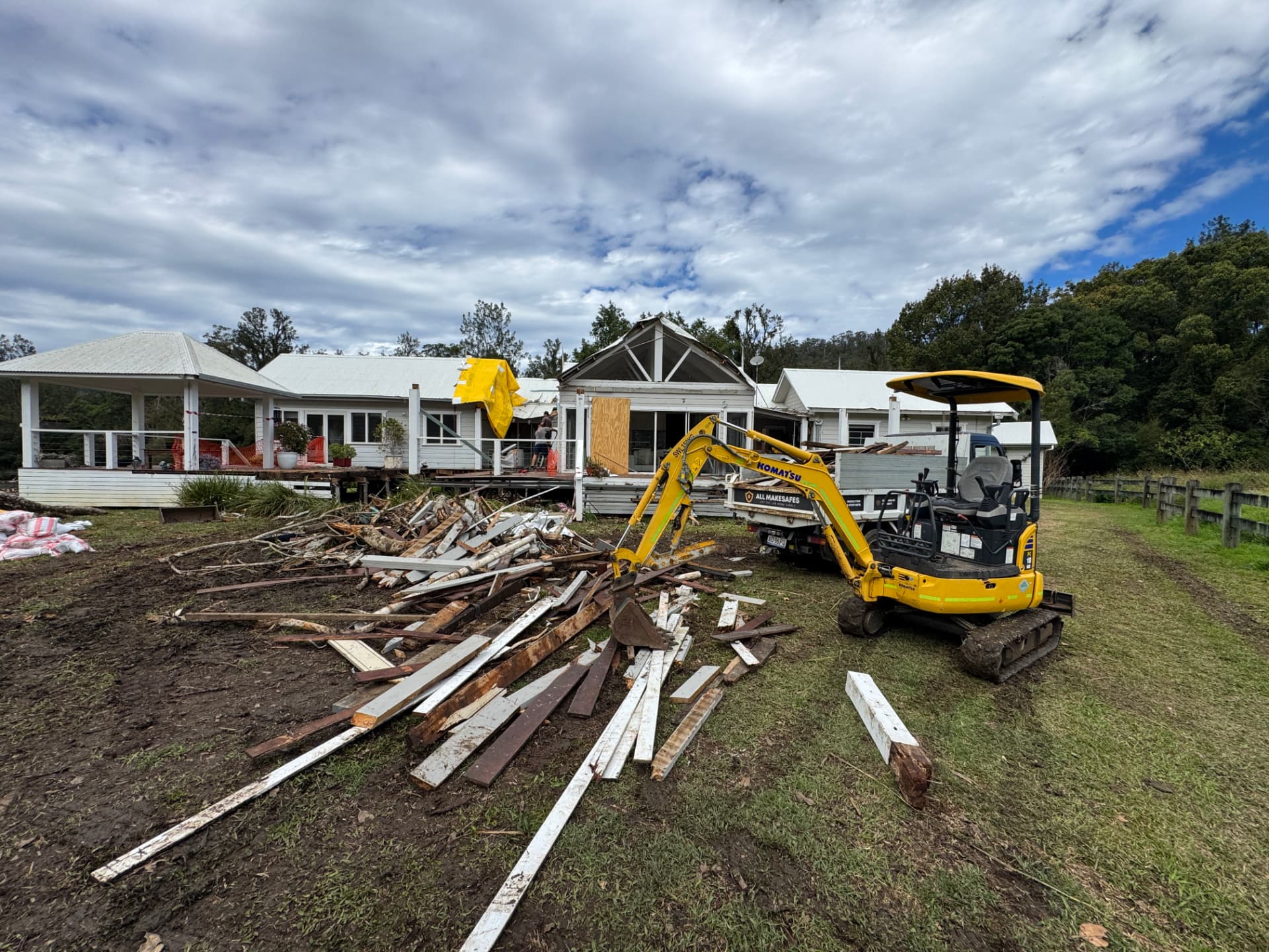 Excavator clearing stripped-out timber and roofing debris beside a storm-damaged Queensland homestead